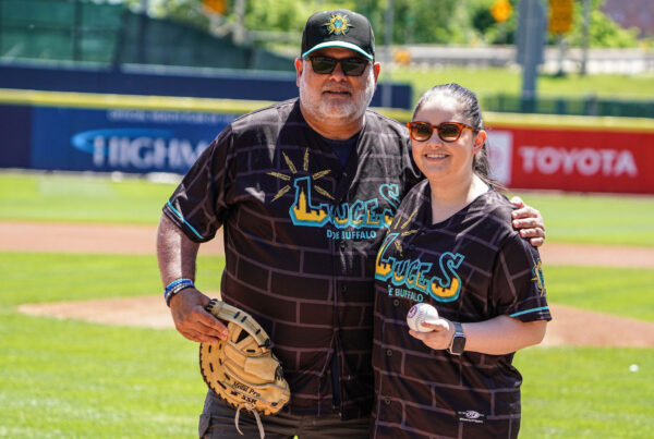 Two people in Luces de Buffalo jerseys holding a baseball and mitt stand together on the field